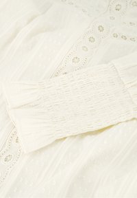 Witte jurk van textuurstof, met kantafwerking, gerimpelde mouwen en kleine stippenpatronen verspreid over de stof. Elegant en delicaat ontwerp.