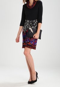 Maglietta nera a maniche lunghe con scollatura a girocollo; gonna ginocchio a fantasia nei toni rosso, blu e nero; tiene una pochette nera con un accento argentato.