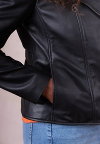 Black leather jacket with a fitted design, visible stitching, and a pocket. Worn over an orange top and blue jeans. Soft texture.