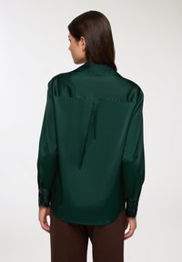 Zielona satynowa koszula o dopasowanym kroju, z długimi rękawami i zakładką z tyłu. Posiada mankiety zapinane na guziki oraz gładką, błyszczącą teksturę.