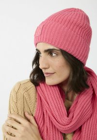 Roze gebreide béret met geribbelde textuur en geborduurd logo, gecombineerd met een roze geribbelde sjaal. Beide accessoires zijn zacht en comfortabel.