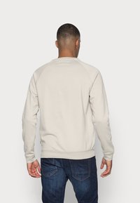 Felpa beige chiaro con collo rotondo e maniche raglan, realizzata in tessuto morbido, abbinata a jeans blu scuro. Nessun motivo o logo visibile.
