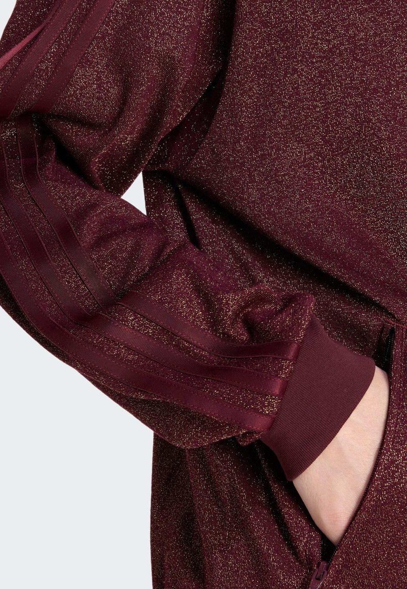 Hand in een glinsterende kastanjebruine jacksleeve met geribbelde manchette en drie satijnen strepen, hand gestoken in een ritszak aan de zijkant.