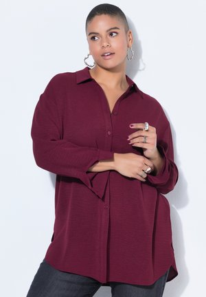 Osoba s krátkými vlasy, oblečená v vínově červené košili na knoflíky, s kruhovými náušnicemi a několika prsteny, pózující na bílém pozadí.