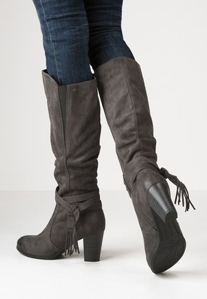 Hohe, dunkelgraue Wildleder-Stiefel mit Absatz und Quasten-Details, getragen über dunkelblauen Skinny-Jeans vor weißem Hintergrund.