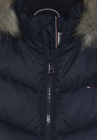 Námorně modrá péřová bunda s kožešinovou kapucí. Obsahuje viditelný zip, diamantově prošívaný design a malý logo detail blízko lemování.