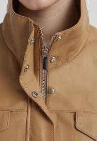 Braune Lederjacke mit hohem Kragen, silbernem Reißverschluss, Druckknopfverschlüssen und sichtbaren Nähten in der Nähe der Taschen.