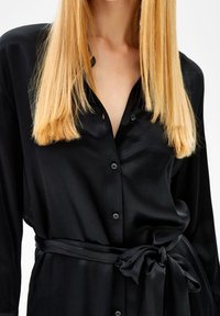 Czarna koszula na guziki o gładkiej fakturze, z paskiem do wiązania w talii, dopasowanym krojem i krótkim kołnierzykiem.