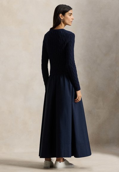 Πλεκτό πουλόβερ σε ναυτικό μπλε με σχέδιο καλώδιο, συνδυασμένο με μια αέρινη, μονόχρωμη φούστα σε ναυτικό μπλε. Το μοντέλο φοράει λευκά αθλητικά παπούτσια, παρουσιάζοντας μια κομψή σιλουέτα.