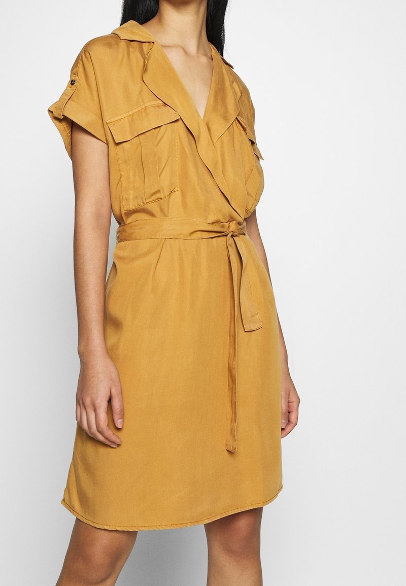 Femme portant une robe courte à manches courtes couleur moutarde, cintrée avec une ceinture, des poches poitrine et un col, sur un fond blanc uni.