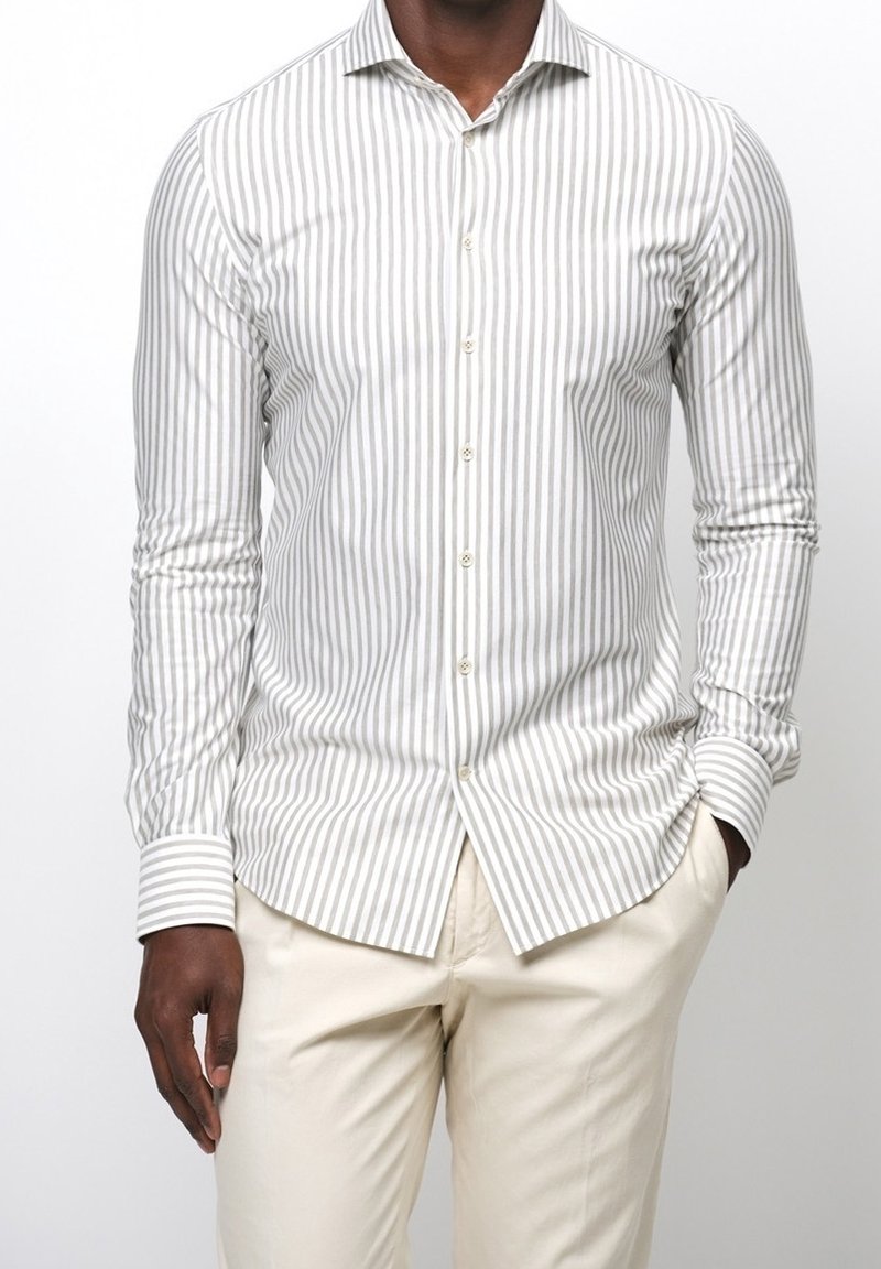 Homme portant une chemise blanche à boutons avec de fines rayures verticales grises et un pantalon beige, une main dans la poche, torse visible.