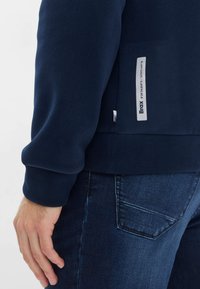 Felpa blu navy realizzata in morbido tessuto con polsini a coste, dotata di un'etichetta laterale e un design posteriore liscio, abbinata a jeans in denim scuro.