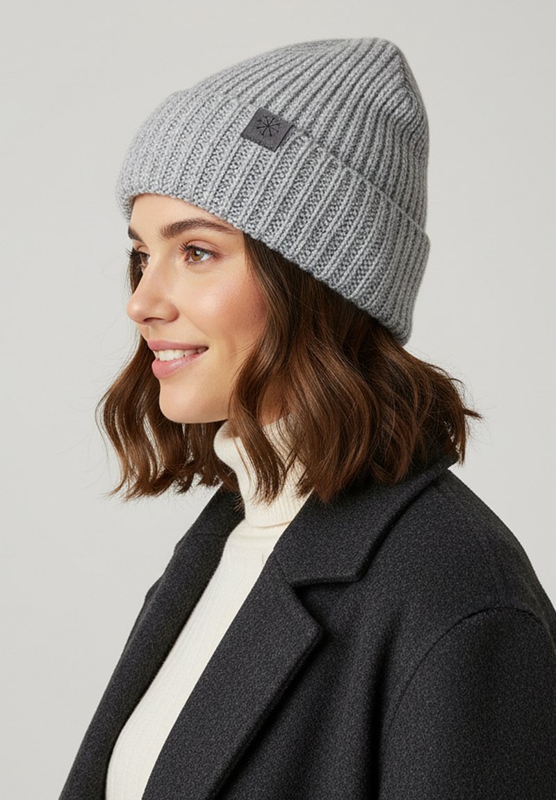 Gray ribbed knit beanie with a folded cuff and a small square logo patch, worn with a cream turtleneck under a dark coat.