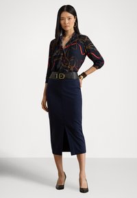 Sieviete stāv, viena roka kabatā, valkājot rakstainu blūzi, tumši zilu midi svārku ar priekšējo šķēlumu, melnu jostu un melnas augstpapēžu kurpes.