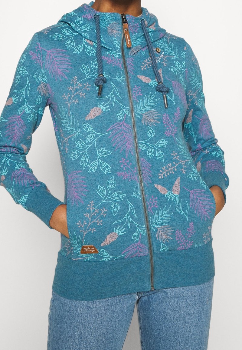Sweat à capuche zippé turquoise avec des motifs floraux en bleu, violet et beige, porté avec un jean bleu clair, mains dans les poches.
