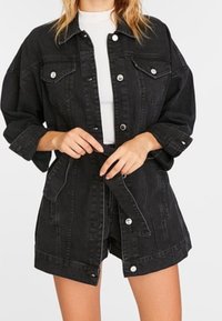Frau trägt eine schwarze Jeansjacke und einen passenden Rocks mit Gürtel über einem weißen Oberteil, die Hände justieren den Gürtel an der Taille.