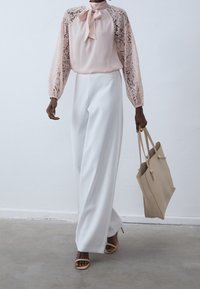 Blouse rose clair avec manches en dentelle et détail nœud, associée à un pantalon blanc à jambes larges. Sac fourre-tout beige et talons nude complètent la tenue.