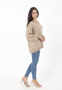 Beige gebreide oversized trui met verlaagde schouders, geribde mouwen en een V-hals, gecombineerd met lichtblauwe skinny jeans en doorzichtige sandalen met hakken.
