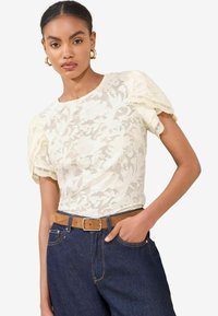 White floral lace top with short puff sleeves, featuring a round neck and lace trim, paired with dark denim high-waisted jeans and a brown belt.