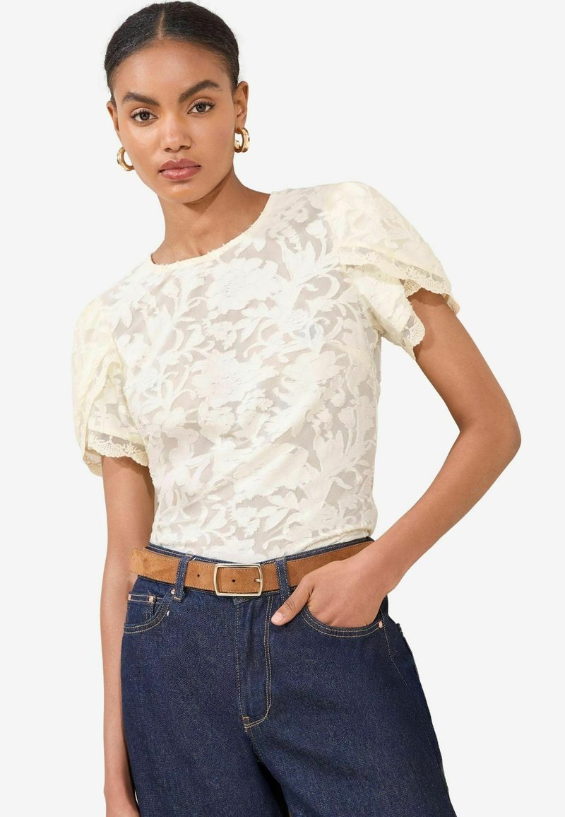 Top in pizzo bianco con maniche a sbuffo corte, caratterizzato da un collo rotondo e bordo in pizzo, abbinato a jeans in denim scuro a vita alta e una cintura marrone.