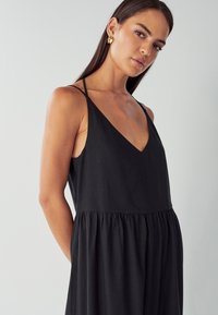 Žena s dlouhými tmavými vlasy, která má na sobě zlaté náušnice a černé šaty bez rukávů s tenkými ramínky, na jednolitém světlém pozadí.