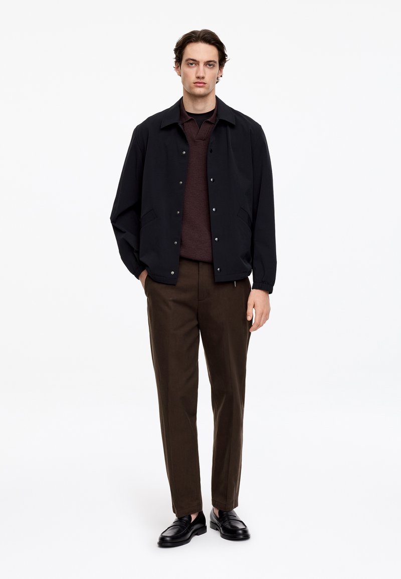 Jeune homme debout, portant une veste noire, un pull marron foncé, un pantalon marron et des mocassins noirs sur fond blanc.