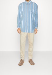 Camisa de botones azul claro con rayas verticales blancas, mangas largas y bolsillo en el pecho, combinada con pantalones beige y zapatos marrones.