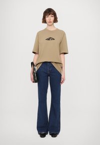 Beige oversized t-shirt met zwart logo, gecombineerd met uitlopende donkerblauwe jeans. Accessoire: kleine gestructureerde zwarte handtas. Schoeisel: zwarte schoenen.