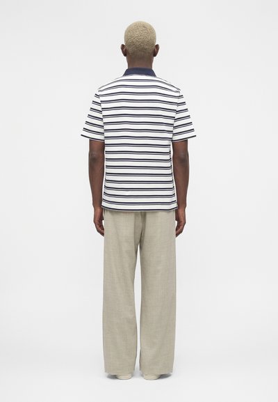 Άτομο με κοντά ξανθά μαλλιά στέκεται με την πλάτη, φοράει λευκό και ναυτικό ριγέ κοντομάνικο πουκάμισο και φαρδιά μπεζ παντελόνια.