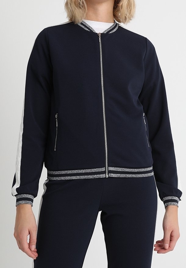 Femme portant une veste zippée bleu marine avec des bordures argentées et des bandes latérales, associée à un pantalon bleu marine assorti.