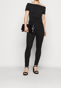 Svart off-shoulder topp med rynkad textur, kombinerad med åtsittande svarta jeans och svarta högklackade sandaler, hållande en svart kviltad clutch.