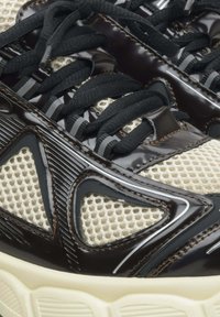 Czarne, błyszczące sneakersy z beżowymi akcentami z siateczki. Posiadają czarne sznurowadła, teksturowane nakładki i masywną białą podeszwę. Model prezentuje skomplikowane detale.