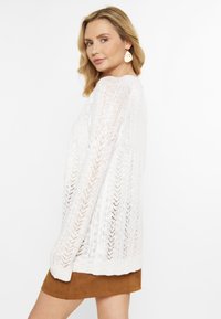 Maglione bianco lavorato a maglia con un motivo a zigzag e vestibilità ampia. Maniche lunghe e orlo rotondo. Indossato con una gonna in suede marrone.