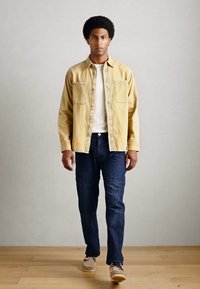 Cămașă din bumbac galben deschis peste un tricou alb, blugi închiși la culoare, pantofi crem; model care merge pe un podea din lemn cu un fundal simplu.