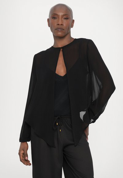 Black sheer blouse with long, flowing sleeves and a rounded hem, featuring a subtle keyhole closure at the front. Paired with black pants.