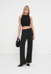 Kvinde iført sort ærmeløs crop top, mørke bredbenede bukser, sorte spidse hæle, bærende på en lille sort håndtaske mod en hvid baggrund.