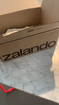 Rudas kartoninis dėžė su dideliu juodu "zalando" logotipu; turinys iš dalies matomas viduje; padėta ant marmuruotomis raštais dangos.