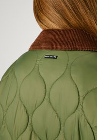 Giacca trapuntata verde con colletto in velluto a coste marrone e un piccolo etichetta logo nera, che presenta una texture liscia e un motivo di cucitura curva.