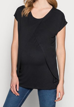 Femme portant un haut de maternité noir à manches courtes avec des panneaux avant superposés et un jean bleu, debout devant un fond uni.