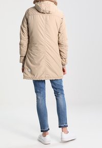 Parka beige isolata con cappuccio staccabile foderato in pelliccia, design trapuntato sulle spalle e un patch con il logo vicino all'orlo. Indossata sopra jeans blu e sneaker bianche.