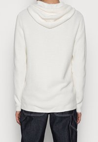 White knitted hoodie with a textured pattern, featuring a drawstring hood, long sleeves, and an elongated silhouette. Paired with dark blue jeans.