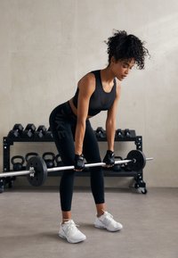 Črna športna majica in leggingi, dvigovanje olimpijske palice s črnimi utežmi. Bele športne čevlje. Notranji fitnes prostor s težkimi utežmi v ozadju.