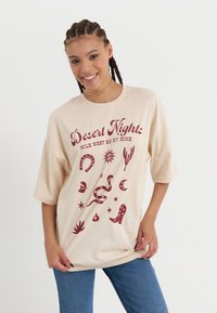 Beige oversized t-shirt met een rode grafische print met symbolen zoals cactussen, sterren en laarzen. Korte mouwen en een ronde halsontwerp.