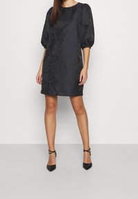 Černé krátké šaty s nafouknutými rukávy, texturovaná látka s jemným vzorem, doplněné černými lodičkami s špičatou špičkou a kotníkovými pásky.