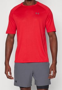 Camiseta atlética roja de manga corta con cuello redondo y logo gris. Llevada con pantalones cortos grises. Textura suave, tela transpirable.