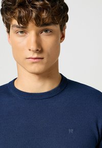 Maglione azzurro navy a collo rotondo lavorato a maglia con scollo a coste e una piccola accentatura del logo sul petto, caratterizzato da una superficie liscia e un design aderente.
