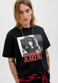 Sort bomulds t-shirt med en kontrasterende film-grafik af en mand med "SCARFACE" tekst i rød. Inkluderer en sølv kædede halskæde og en tatoveret arm.