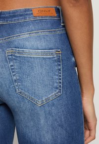 Primer plano de un bolsillo trasero de unos vaqueros de mezclilla azul con una etiqueta de cuero marrón que dice "ONLY" en la cadera de una persona, con el brazo visible.