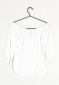 Blusa bianca off-the-shoulder con scollatura smock, maniche a tre quarti e dettagli annodati sui polsini, appesa a un gancio nero.