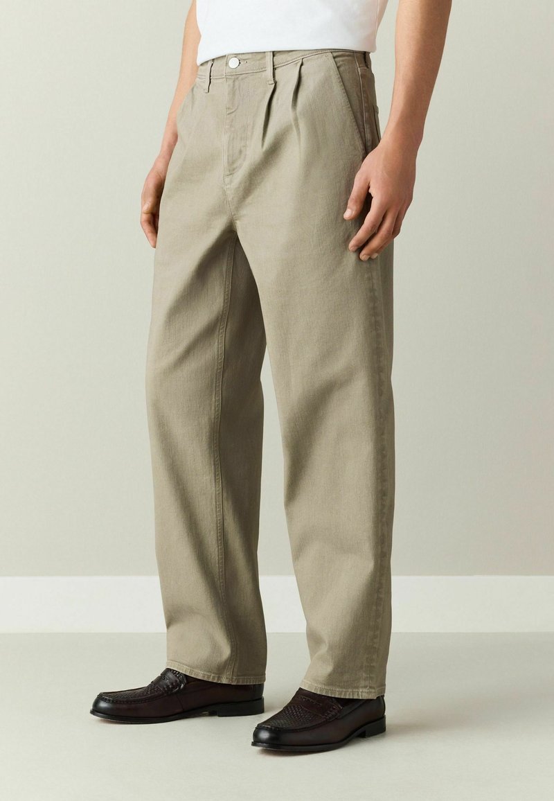 Homme portant un pantalon beige coupe droite, une chemise blanche et des mocassins marron foncé, debout devant un fond neutre.